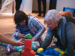 Girl with glasses and dark hair sits on the floor and plays with blocks with older woman