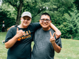 Two teenage boys pose for the camera with one arm around each other. One boy is shows a peace sign and has glasses. The other boy is wearing a hat and has his thumb and pinky out.